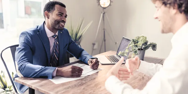 Two men sitting at a table in an office, engaged in conversation. One man wears a blue suit, and a laptop and plant are on the table.