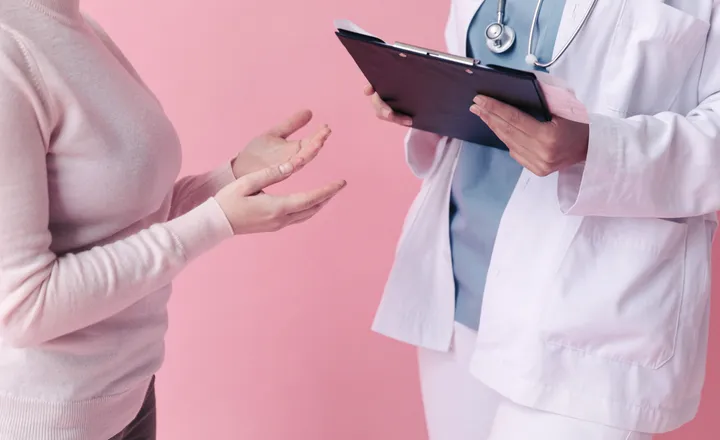 A doctor holding a clipboard talks with a patient gesturing, against a pink background.