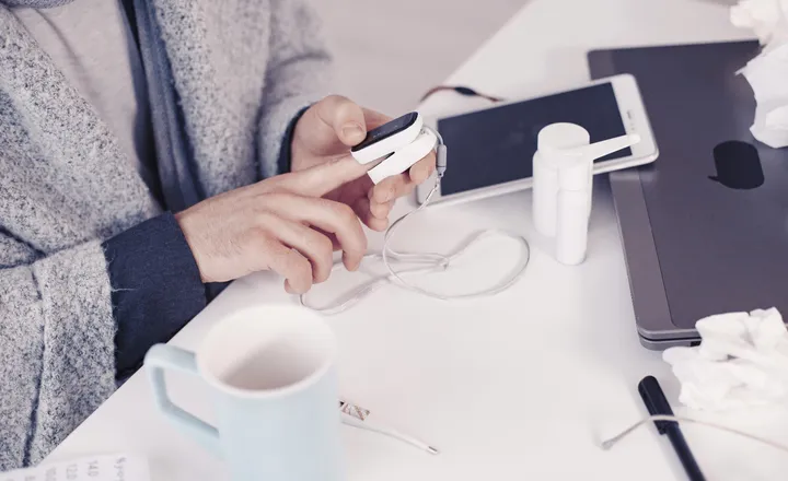 Person using a fingertip pulse oximeter at a desk with a mug, tissues, nasal spray, thermometer, and tablet.