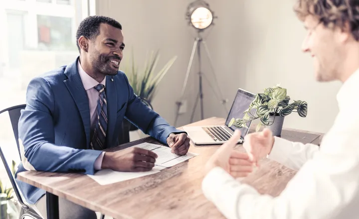Two men sitting at a table in an office, engaged in conversation. One man wears a blue suit, and a laptop and plant are on the table.