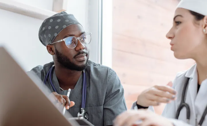 Two healthcare professionals in discussion, sitting indoors near a window, wearing scrubs and stethoscopes, with a laptop in front.