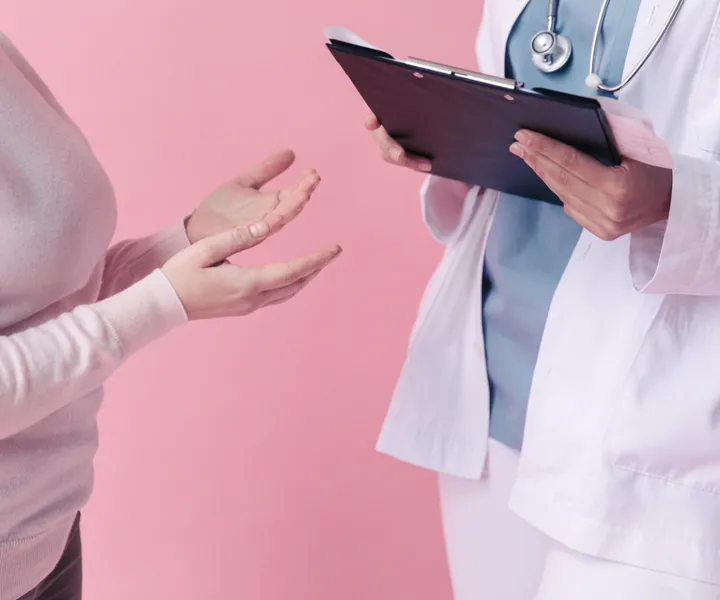 A doctor holding a clipboard talks with a patient gesturing, against a pink background.