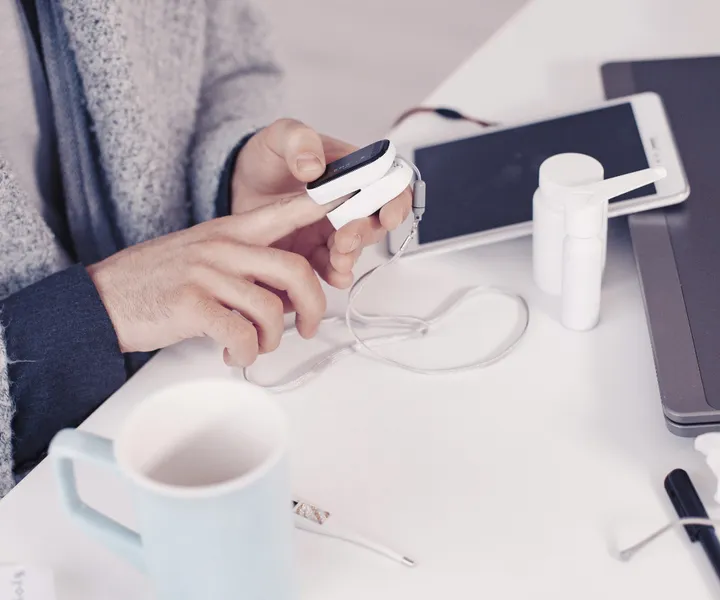 Person using a fingertip pulse oximeter at a desk with a mug, tissues, nasal spray, thermometer, and tablet.