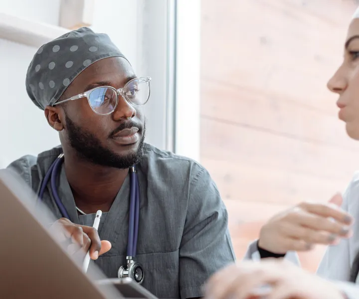 Two healthcare professionals in discussion, sitting indoors near a window, wearing scrubs and stethoscopes, with a laptop in front.